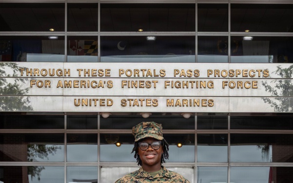 A United States Marine in woodland MARPAT camouflage uniform stands proudly in front of a building bearing the inscription 'Through These Portals Pass Prospects For America's Finest Fighting Force - United States Marines.' The Marine is holding an official document, likely a certificate or enlistment/promotion order with a red border, and is smiling at the camera. The setting appears to be a Marine Corps recruiting station or administrative building.