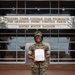 A United States Marine in woodland MARPAT camouflage uniform stands proudly in front of a building bearing the inscription 'Through These Portals Pass Prospects For America's Finest Fighting Force - United States Marines.' The Marine is holding an official document, likely a certificate or enlistment/promotion order with a red border, and is smiling at the camera. The setting appears to be a Marine Corps recruiting station or administrative building.