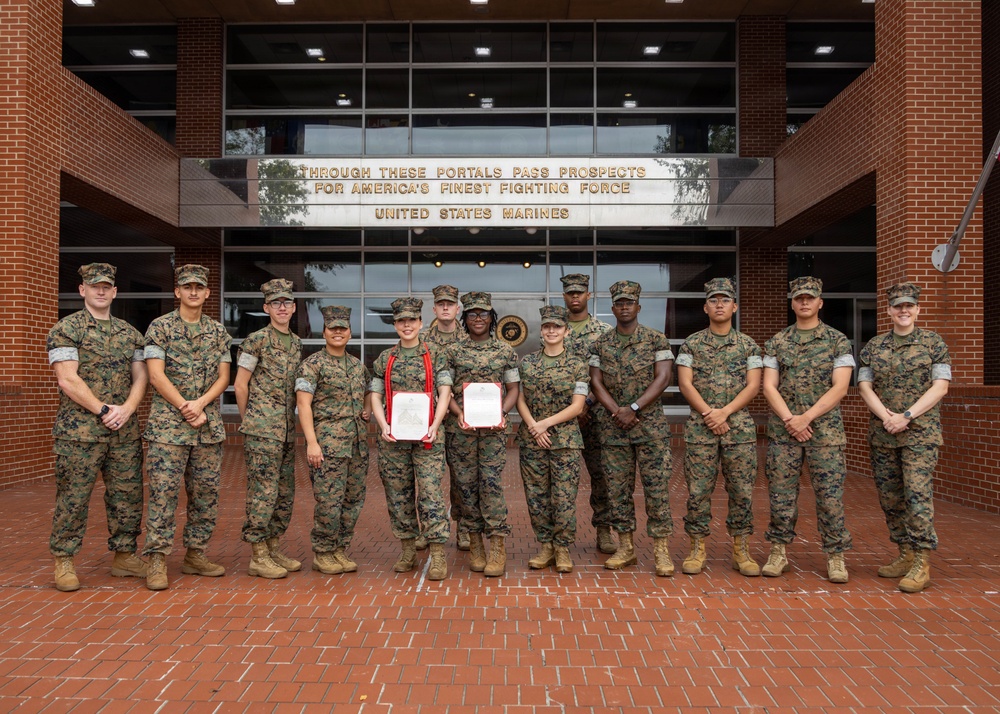 A group of approximately 13 United States Marines in woodland MARPAT camouflage uniforms pose for a formal group photograph in front of a brick building. Two Marines in the center are holding framed certificates or awards with red ribbons, suggesting a recognition or award ceremony. The building's entrance displays the iconic inscription: 'Through These Portals Pass Prospects for America's Finest Fighting Force - United States Marines.'