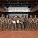 A group of approximately 13 United States Marines in woodland MARPAT camouflage uniforms pose for a formal group photograph in front of a brick building. Two Marines in the center are holding framed certificates or awards with red ribbons, suggesting a recognition or award ceremony. The building's entrance displays the iconic inscription: 'Through These Portals Pass Prospects for America's Finest Fighting Force - United States Marines.'