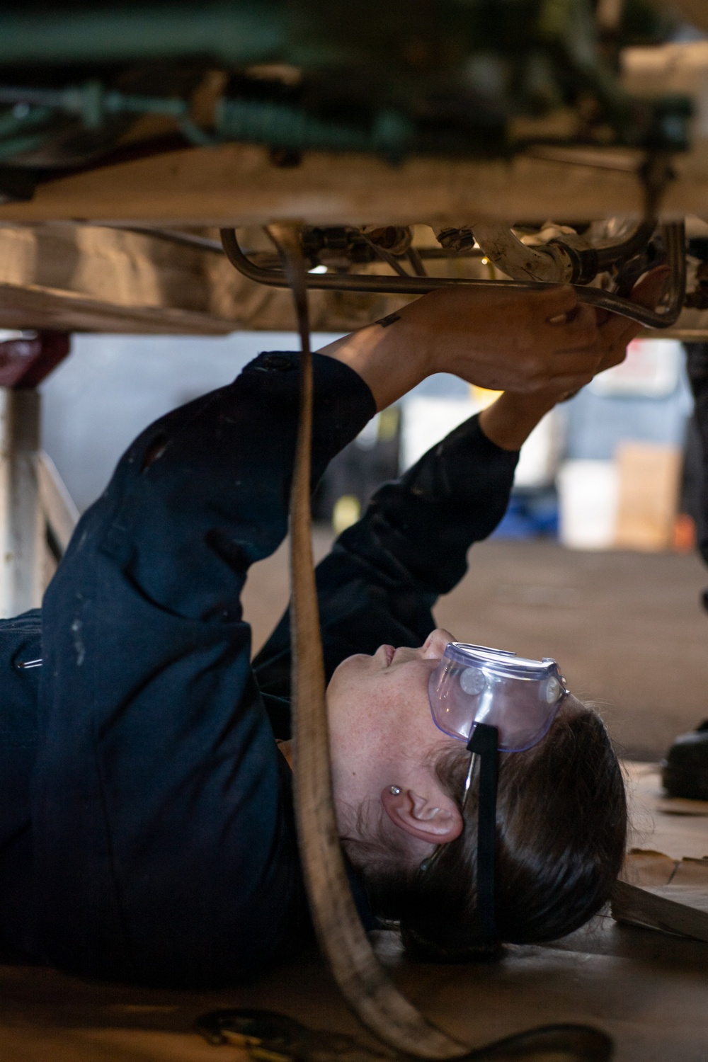 A mechanic is lying on their back on the floor beneath a vehicle, working on the undercarriage components. The person is wearing protective safety goggles and dark work coveralls, reaching upward with both hands to manipulate parts on the vehicle's underside. The setting appears to be an automotive repair shop or garage.