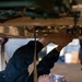 A mechanic is lying on their back on the floor beneath a vehicle, working on the undercarriage components. The person is wearing protective safety goggles and dark work coveralls, reaching upward with both hands to manipulate parts on the vehicle's underside. The setting appears to be an automotive repair shop or garage.