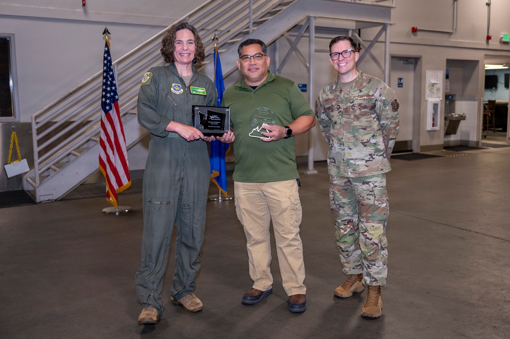 Three individuals pose together in what appears to be a military hangar or facility during an awards ceremony. The person in the center is holding two awards — a plaque and a glass trophy — flanked by a person in an olive green flight suit on the left and a person in OCP (Operational Camouflage Pattern) military uniform on the right. An American flag and a blue flag (likely Air Force) are visible in the background.