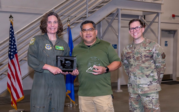 Three individuals pose together in what appears to be a military hangar or facility during an awards ceremony. The person in the center is holding two awards — a plaque and a glass trophy — flanked by a person in an olive green flight suit on the left and a person in OCP (Operational Camouflage Pattern) military uniform on the right. An American flag and a blue flag (likely Air Force) are visible in the background.