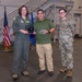 Three individuals pose together in what appears to be a military hangar or facility during an awards ceremony. The person in the center is holding two awards — a plaque and a glass trophy — flanked by a person in an olive green flight suit on the left and a person in OCP (Operational Camouflage Pattern) military uniform on the right. An American flag and a blue flag (likely Air Force) are visible in the background.