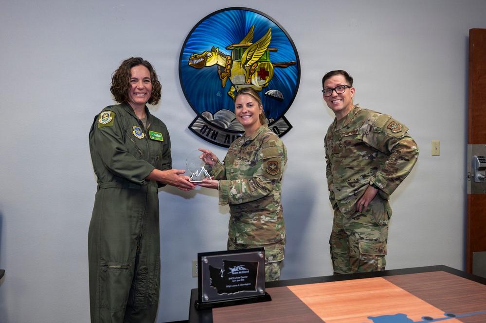 Three military personnel are posing for a photo during an award presentation ceremony. Two individuals are jointly holding a crystal/glass trophy award, while a third stands nearby smiling. A plaque on the table in the foreground reads 'Team McChord' and references a quarterly award.