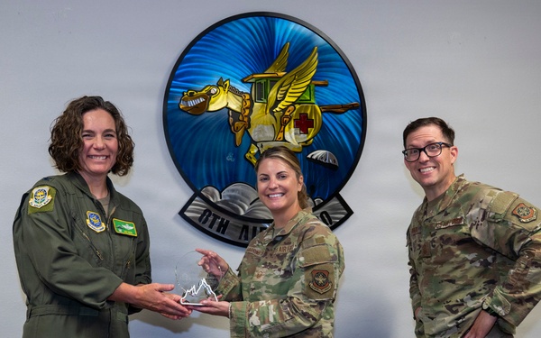 Three military personnel are posing for a photo during an award presentation ceremony. Two individuals are jointly holding a crystal/glass trophy award, while a third stands nearby smiling. A plaque on the table in the foreground reads 'Team McChord' and references a quarterly award.