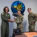 Three military personnel are posing for a photo during an award presentation ceremony. Two individuals are jointly holding a crystal/glass trophy award, while a third stands nearby smiling. A plaque on the table in the foreground reads 'Team McChord' and references a quarterly award.