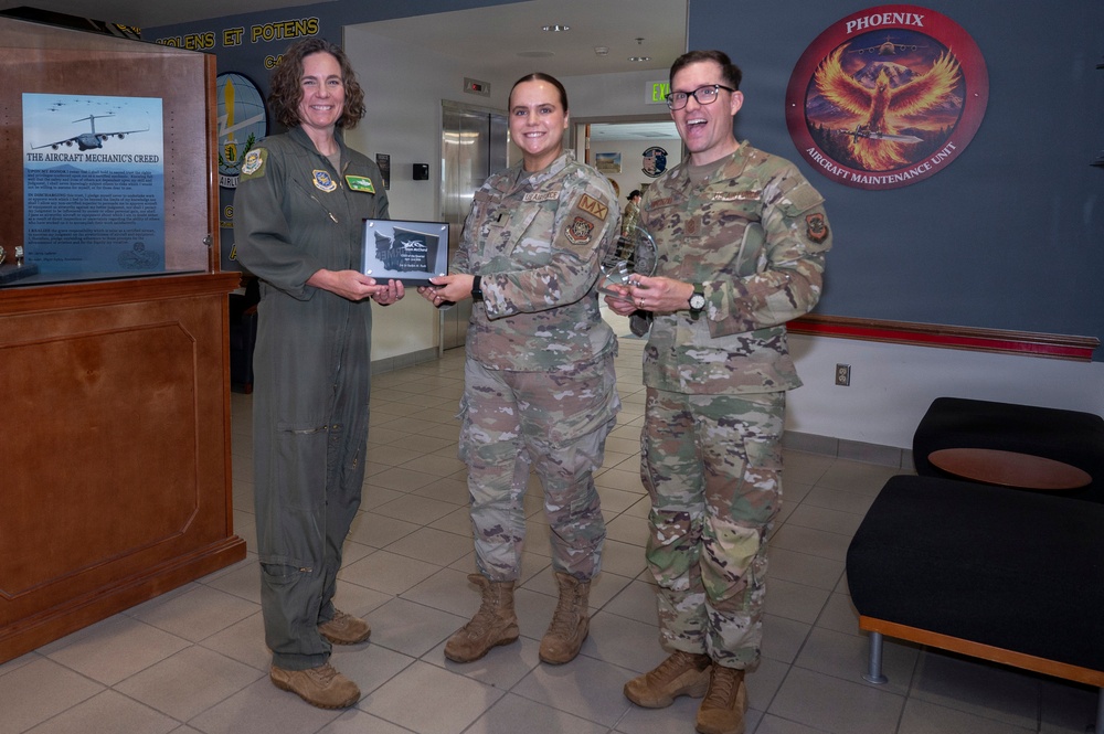 Three U.S. Air Force personnel are posing for a photo in what appears to be a military facility lobby. Two individuals in OCP (Operational Camouflage Pattern) uniforms and one in an olive green flight suit are holding awards, including a plaque and a crystal trophy. The setting appears to be an award or recognition ceremony at an Aircraft Maintenance Unit.