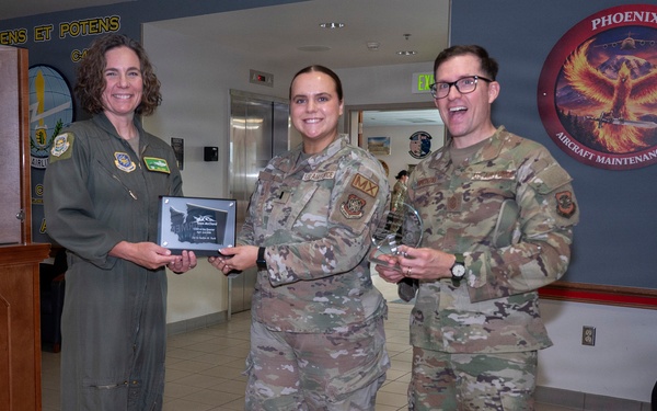 Three U.S. Air Force personnel are posing for a photo in what appears to be a military facility lobby. Two individuals in OCP (Operational Camouflage Pattern) uniforms and one in an olive green flight suit are holding awards, including a plaque and a crystal trophy. The setting appears to be an award or recognition ceremony at an Aircraft Maintenance Unit.
