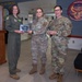 Three U.S. Air Force personnel are posing for a photo in what appears to be a military facility lobby. Two individuals in OCP (Operational Camouflage Pattern) uniforms and one in an olive green flight suit are holding awards, including a plaque and a crystal trophy. The setting appears to be an award or recognition ceremony at an Aircraft Maintenance Unit.