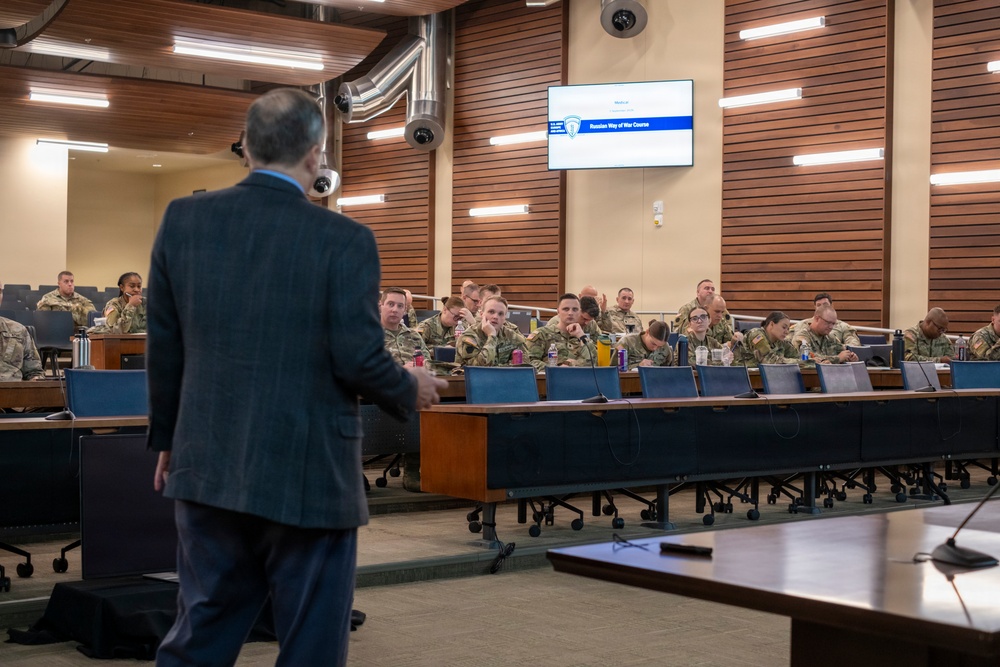 A civilian instructor in a dark suit is presenting a 'Russian Way of War Course' to a room full of military personnel in camouflage uniforms. The setting appears to be a modern military education facility or conference room with tiered seating. The students are seated at long wooden tables and appear attentive to the lecture.