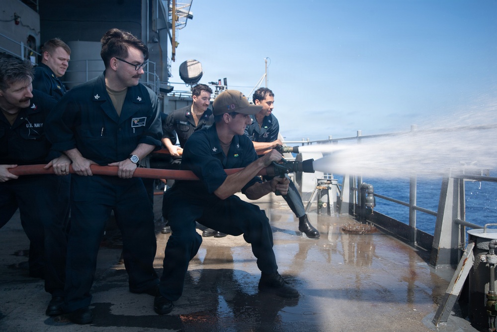 A group of U.S. Navy sailors are conducting a firefighting hose drill on the deck of a naval vessel at sea. Several personnel in dark navy uniforms are gripping a red fire hose while one sailor in a crouched position operates the nozzle, directing a powerful stream of water. The wet deck and open ocean visible in the background confirm this is an active at-sea damage control training exercise.