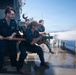A group of U.S. Navy sailors are conducting a firefighting hose drill on the deck of a naval vessel at sea. Several personnel in dark navy uniforms are gripping a red fire hose while one sailor in a crouched position operates the nozzle, directing a powerful stream of water. The wet deck and open ocean visible in the background confirm this is an active at-sea damage control training exercise.