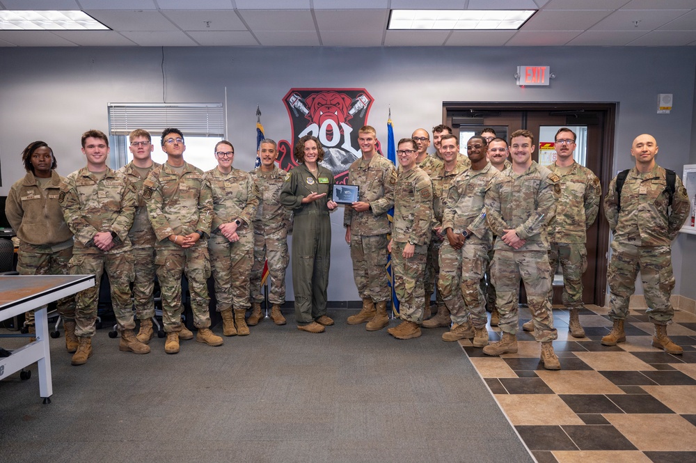 A group of approximately 18-20 military personnel pose for a formal group photo in what appears to be a unit operations or briefing room. One individual in the center, wearing an olive green flight suit, is holding a plaque or award, suggesting a recognition or award ceremony. A unit emblem featuring a bulldog mascot with the letters 'COL' is prominently displayed on the wall behind the group.