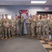 A group of approximately 18-20 military personnel pose for a formal group photo in what appears to be a unit operations or briefing room. One individual in the center, wearing an olive green flight suit, is holding a plaque or award, suggesting a recognition or award ceremony. A unit emblem featuring a bulldog mascot with the letters 'COL' is prominently displayed on the wall behind the group.