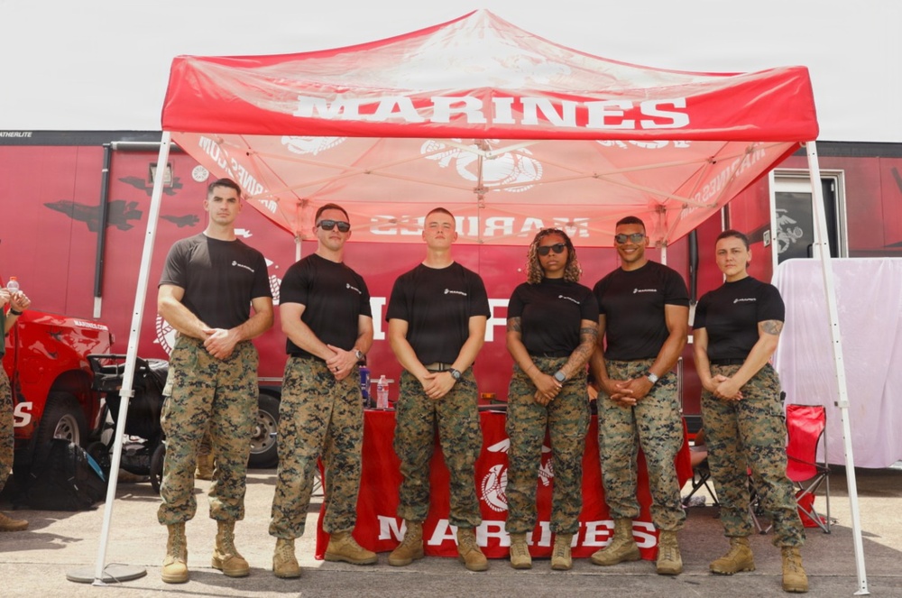 Six U.S. Marines in uniform stand together under a red 'Marines' branded canopy tent at what appears to be a public outreach or recruiting event. They are all wearing matching black t-shirts with Marine Corps branding and digital camouflage (MARPAT) trousers with tan combat boots. A Marines-branded trailer and promotional table are visible in the background.