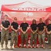 Six U.S. Marines in uniform stand together under a red 'Marines' branded canopy tent at what appears to be a public outreach or recruiting event. They are all wearing matching black t-shirts with Marine Corps branding and digital camouflage (MARPAT) trousers with tan combat boots. A Marines-branded trailer and promotional table are visible in the background.