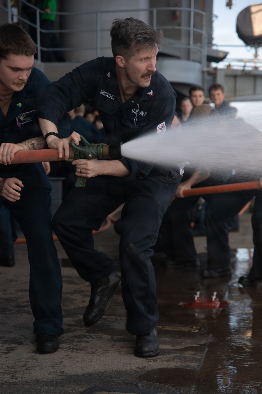 Two U.S. Navy sailors in dark blue uniforms are operating a fire hose on what appears to be a ship's deck during a firefighting drill or training exercise. The central sailor, identified by name tape as 'RHOADES,' is bracing against the water pressure while directing a powerful stream from the nozzle. A group of observers can be seen in the background, suggesting this is a demonstration or training scenario.