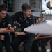Two U.S. Navy sailors in dark blue uniforms are operating a fire hose on what appears to be a ship's deck during a firefighting drill or training exercise. The central sailor, identified by name tape as 'RHOADES,' is bracing against the water pressure while directing a powerful stream from the nozzle. A group of observers can be seen in the background, suggesting this is a demonstration or training scenario.