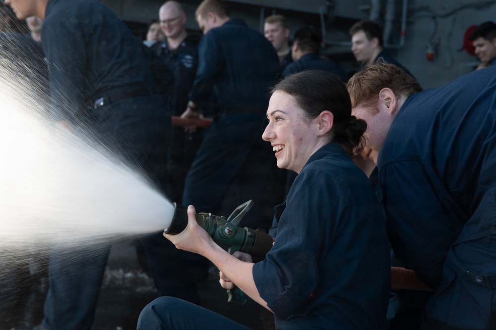 A group of individuals wearing matching dark navy blue uniforms are engaged in what appears to be a firefighting or damage control training exercise. A young woman in the foreground is smiling and operating a fire hose nozzle, directing a powerful stream of water or foam to the left. Several other uniformed personnel are visible in the background observing or participating in the drill.