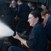 A group of individuals wearing matching dark navy blue uniforms are engaged in what appears to be a firefighting or damage control training exercise. A young woman in the foreground is smiling and operating a fire hose nozzle, directing a powerful stream of water or foam to the left. Several other uniformed personnel are visible in the background observing or participating in the drill.