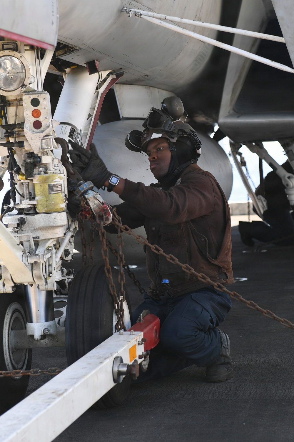 A naval aviation deck crew member is working on the landing gear assembly of a military aircraft, likely on an aircraft carrier flight deck. The individual is kneeling and handling chains and mechanical components near the nose gear of the aircraft. They are wearing standard flight deck personal protective equipment including a helmet, goggles, hearing protection, and gloves.