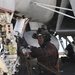 A naval aviation deck crew member is working on the landing gear assembly of a military aircraft, likely on an aircraft carrier flight deck. The individual is kneeling and handling chains and mechanical components near the nose gear of the aircraft. They are wearing standard flight deck personal protective equipment including a helmet, goggles, hearing protection, and gloves.