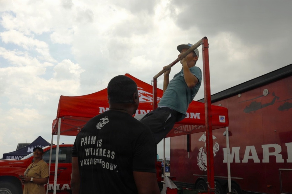 A person in a light blue shirt and cap is performing pull-ups on a red pull-up bar station at what appears to be a U.S. Marines recruiting event or exhibition. A man in a black t-shirt reading 'Pain is Weakness Leaving the Body' — a well-known Marine Corps motto — stands nearby, appearing to spot or count repetitions. The setting includes a red Marines canopy tent, a large Marines vehicle or trailer, and a Navy tent visible in the background.