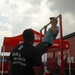 A person in a light blue shirt and cap is performing pull-ups on a red pull-up bar station at what appears to be a U.S. Marines recruiting event or exhibition. A man in a black t-shirt reading 'Pain is Weakness Leaving the Body' — a well-known Marine Corps motto — stands nearby, appearing to spot or count repetitions. The setting includes a red Marines canopy tent, a large Marines vehicle or trailer, and a Navy tent visible in the background.