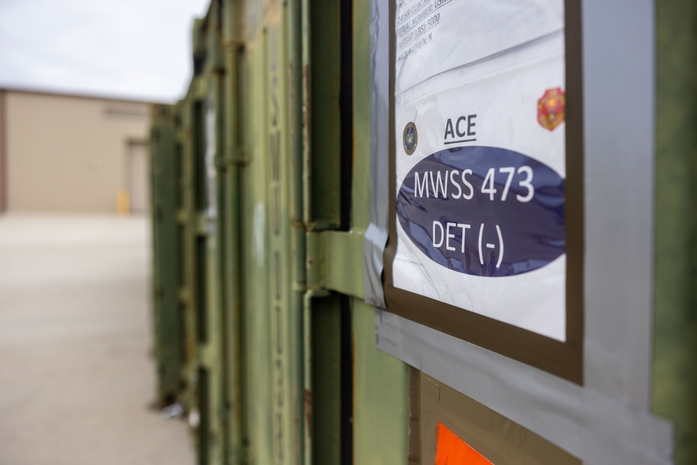 The image shows a row of olive-green military storage containers or conex boxes lined up outdoors near a building. The foreground container has a label affixed to it reading 'ACE' and 'MWSS 473 DET (-)' along with what appear to be military unit insignia patches or stickers. The setting appears to be a military installation or staging area.