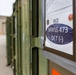 The image shows a row of olive-green military storage containers or conex boxes lined up outdoors near a building. The foreground container has a label affixed to it reading 'ACE' and 'MWSS 473 DET (-)' along with what appear to be military unit insignia patches or stickers. The setting appears to be a military installation or staging area.