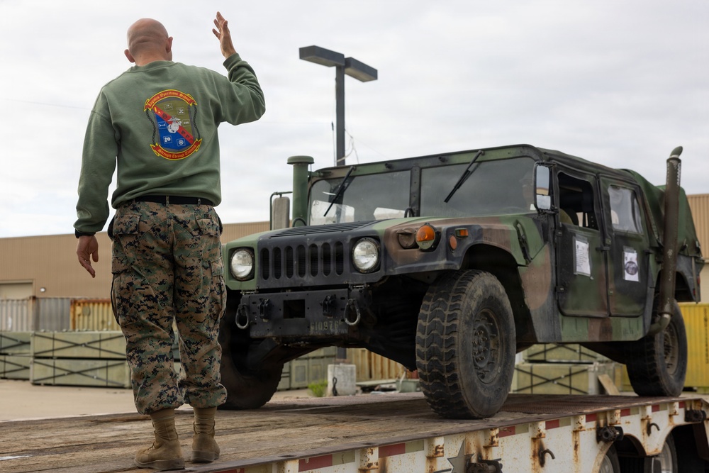 A U.S. Marine in digital camouflage pants and an olive green unit sweatshirt is directing or guiding a military HMMWV (Humvee) that is loaded onto a flatbed trailer. The Marine has his right hand raised, likely signaling to the vehicle operator. The setting appears to be a military installation or logistics yard.