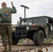 A U.S. Marine in digital camouflage pants and an olive green unit sweatshirt is directing or guiding a military HMMWV (Humvee) that is loaded onto a flatbed trailer. The Marine has his right hand raised, likely signaling to the vehicle operator. The setting appears to be a military installation or logistics yard.