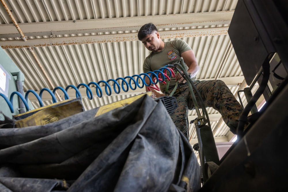 A U.S. Marine in olive drab t-shirt and digital camouflage (MARPAT) trousers is working with coiled air hoses (blue and red/pink) in what appears to be a maintenance bay or motor pool facility. The photo is taken from a low angle, looking upward, giving a dramatic perspective of the service member handling equipment. The setting appears to be an industrial or military maintenance structure with a corrugated metal ceiling showing signs of rust.