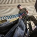 A U.S. Marine in olive drab t-shirt and digital camouflage (MARPAT) trousers is working with coiled air hoses (blue and red/pink) in what appears to be a maintenance bay or motor pool facility. The photo is taken from a low angle, looking upward, giving a dramatic perspective of the service member handling equipment. The setting appears to be an industrial or military maintenance structure with a corrugated metal ceiling showing signs of rust.
