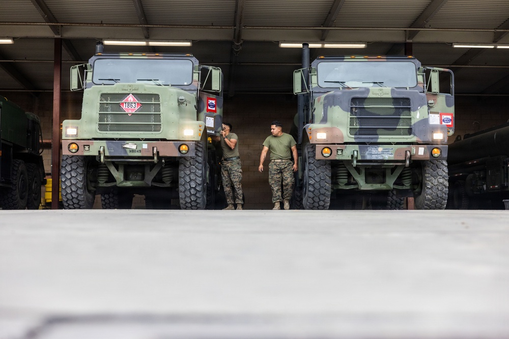 Two military personnel in camouflage uniforms stand between two large olive-drab military trucks inside a covered vehicle bay or motor pool. The trucks appear to be heavy tactical vehicles, likely used for fuel or cargo transport, identifiable by their large grille guards and hazmat placards. The setting suggests a routine maintenance or pre-mission inspection activity at a military installation.