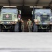 Two military personnel in camouflage uniforms stand between two large olive-drab military trucks inside a covered vehicle bay or motor pool. The trucks appear to be heavy tactical vehicles, likely used for fuel or cargo transport, identifiable by their large grille guards and hazmat placards. The setting suggests a routine maintenance or pre-mission inspection activity at a military installation.