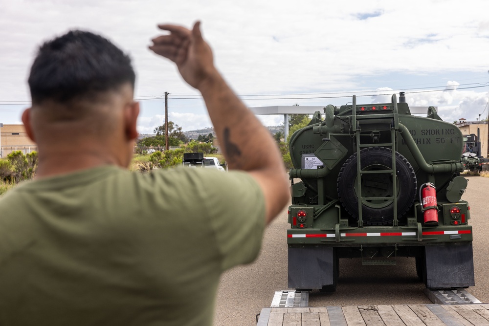 A person in an olive green t-shirt is directing a large military tanker truck, visible from behind with one arm raised in a guiding gesture. The green military vehicle bears 'USMC' markings and a 'NO SMOKING' warning, consistent with a fuel or water tanker. The scene appears to take place at a military installation or staging area.