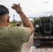 A person in an olive green t-shirt is directing a large military tanker truck, visible from behind with one arm raised in a guiding gesture. The green military vehicle bears 'USMC' markings and a 'NO SMOKING' warning, consistent with a fuel or water tanker. The scene appears to take place at a military installation or staging area.
