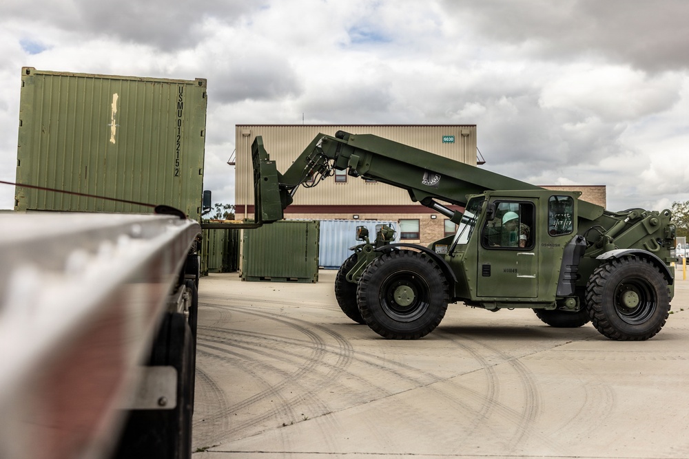 A military olive-drab painted rough terrain container handler (RTCH) is maneuvering in a paved logistics yard, apparently loading or unloading a green military shipping container marked 'USMU0122152'. The scene appears to be at a military installation or depot, with a warehouse building visible in the background. Tire tracks on the concrete surface indicate active vehicle movement in the area.