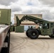 A military olive-drab painted rough terrain container handler (RTCH) is maneuvering in a paved logistics yard, apparently loading or unloading a green military shipping container marked 'USMU0122152'. The scene appears to be at a military installation or depot, with a warehouse building visible in the background. Tire tracks on the concrete surface indicate active vehicle movement in the area.
