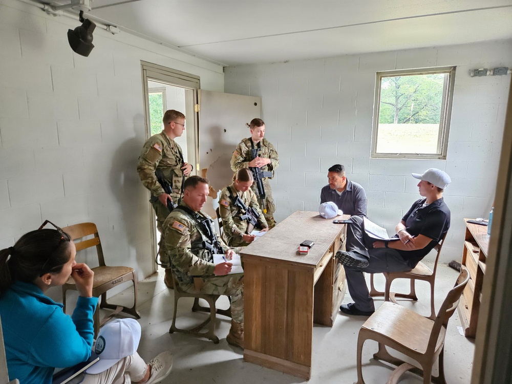 A group of U.S. Army soldiers in OCP (Operational Camouflage Pattern) uniforms are engaged in what appears to be a training exercise or briefing inside a sparse, cinder-block room. Several soldiers are armed with rifles and are interacting with civilians seated at and around a wooden desk. The setting resembles a military training facility used for scenario-based exercises.