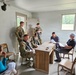 A group of U.S. Army soldiers in OCP (Operational Camouflage Pattern) uniforms are engaged in what appears to be a training exercise or briefing inside a sparse, cinder-block room. Several soldiers are armed with rifles and are interacting with civilians seated at and around a wooden desk. The setting resembles a military training facility used for scenario-based exercises.