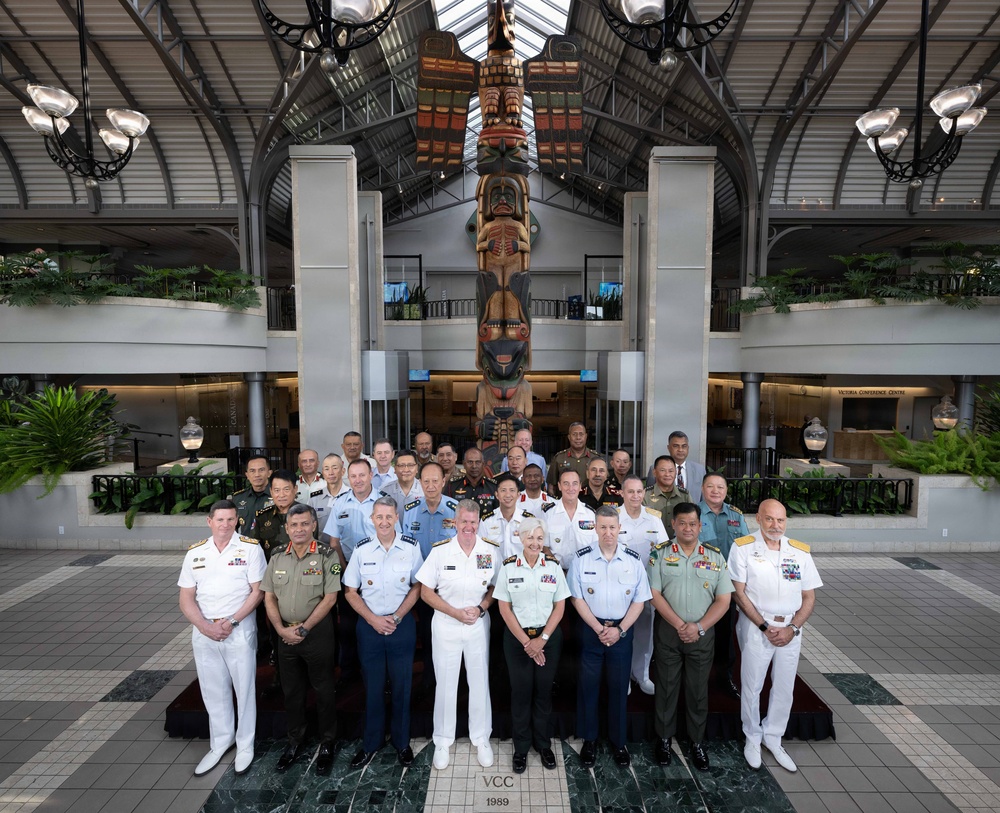 A formal group photograph of approximately 25-30 military officers in various national uniforms posing inside an atrium building. The setting features a prominent totem pole as a backdrop, along with a glass skylight ceiling and indoor tropical plants. A plaque reading 'VCC 1989' is visible at the base of the display, suggesting this is the Victoria Conference Centre.