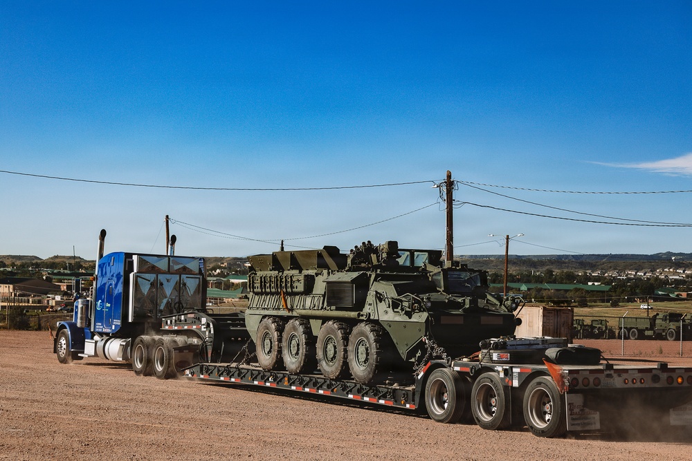 A blue commercial semi-truck is transporting a large olive-green military wheeled armored vehicle, consistent with a Stryker-type 8x8 armored personnel carrier, on a flatbed lowboy trailer. The vehicle is secured with chains on a dirt/gravel surface in what appears to be a military installation or staging area. Additional military vehicles are visible in the background.