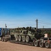 A blue commercial semi-truck is transporting a large olive-green military wheeled armored vehicle, consistent with a Stryker-type 8x8 armored personnel carrier, on a flatbed lowboy trailer. The vehicle is secured with chains on a dirt/gravel surface in what appears to be a military installation or staging area. Additional military vehicles are visible in the background.