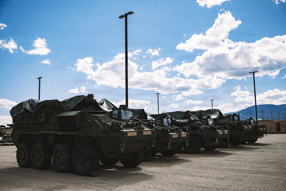A row of military armored wheeled vehicles, consistent with M1126 Stryker Infantry Carrier Vehicles, are parked in a large paved lot at what appears to be a military installation. The vehicles are covered with dark tarpaulins and painted in olive drab camouflage. Mountains and a partly cloudy sky are visible in the background.