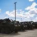 A row of military armored wheeled vehicles, consistent with M1126 Stryker Infantry Carrier Vehicles, are parked in a large paved lot at what appears to be a military installation. The vehicles are covered with dark tarpaulins and painted in olive drab camouflage. Mountains and a partly cloudy sky are visible in the background.