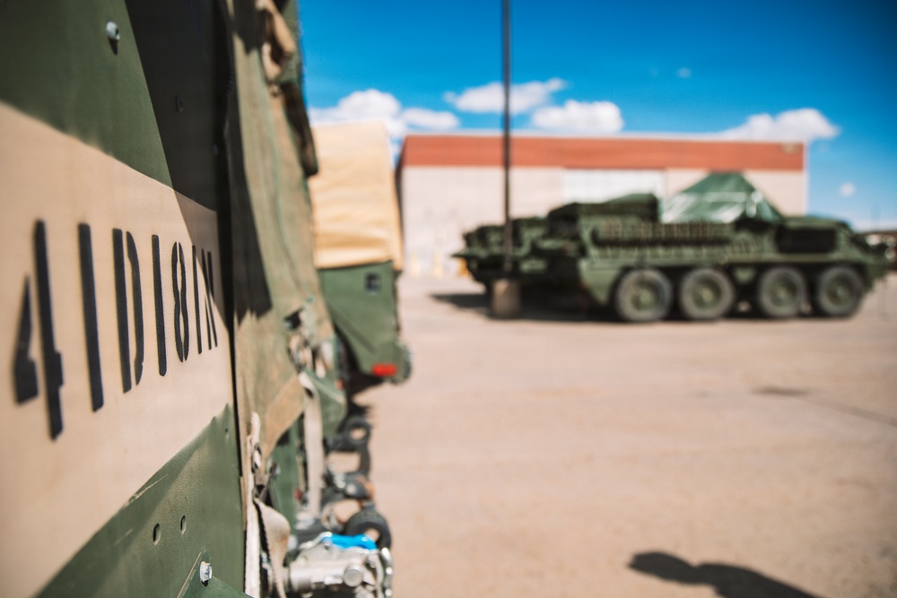 The image shows military vehicles in what appears to be a base or staging area under a clear blue sky. In the foreground, a close-up of a military vehicle panel displays stenciled markings reading '4ID10N' in black lettering on a two-tone green and tan camouflage surface. In the background, a green 8-wheeled armored vehicle, consistent with a Stryker-type APC, is parked near a building.